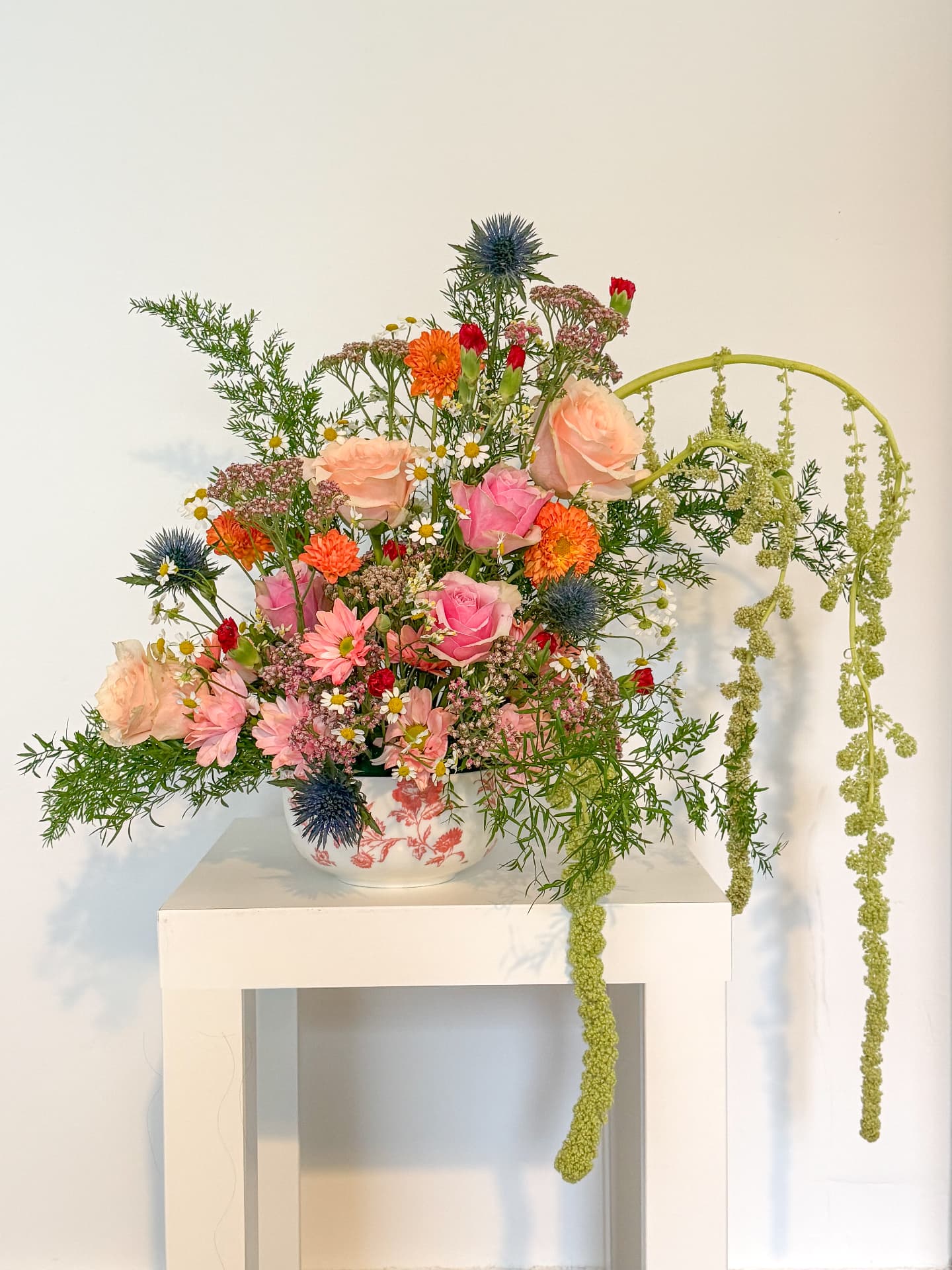 A lush seasonal arrangement of peach and pink roses, orange dahlias, blue thistle and trailing green amaranthus in a floral china vase
